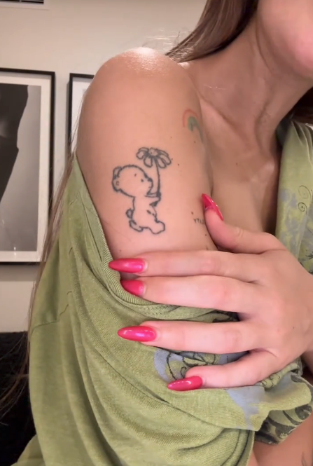 Person showing an limb  tattoo of a playful cartoon dinosaur holding a flower. Wearing a casual, greenish  off-shoulder apical  and agelong  pinkish  nails