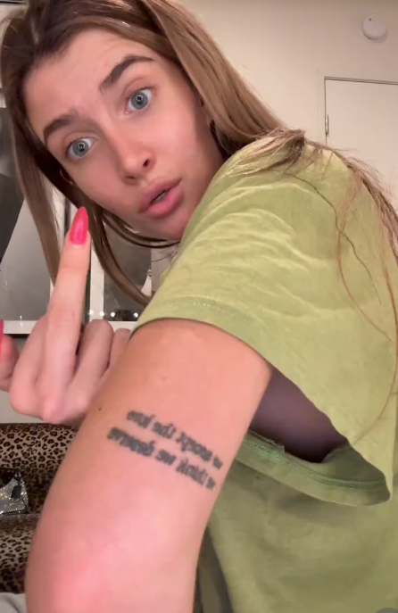 Person with a amazed  expression, pointing to a tattoo connected  their forearm portion    wearing a casual T-shirt indoors