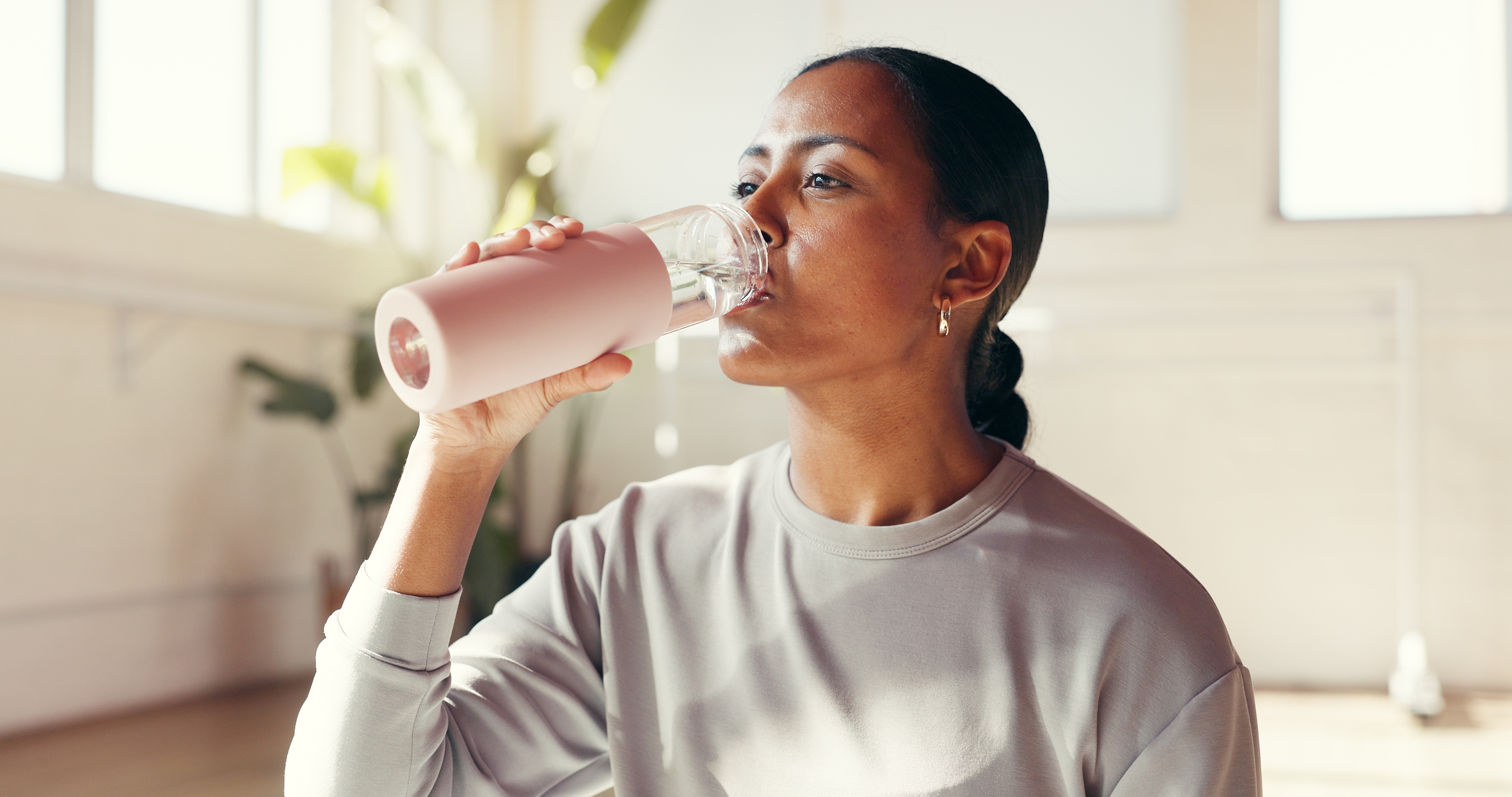 Person drinking from a reusable h2o  vessel  indoors, wearing a sweatshirt, with plants and ample  windows successful  the background