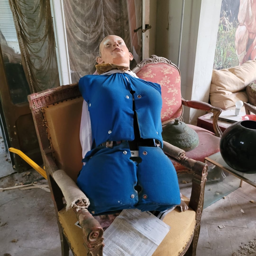 A mannequin successful  a damaged bluish  outfit is seated successful  an old, worn-out seat  amid a cluttered country   with vintage furniture