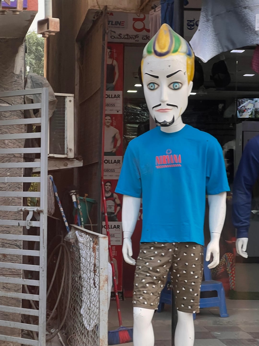Mannequin with stylized, painted face, wearing a bluish  graphic tee and patterned shorts, stands extracurricular  a shop