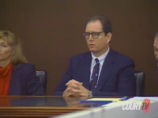 A person in a courtroom sits with hands clasped, wearing a suit and glasses, next to others at a table