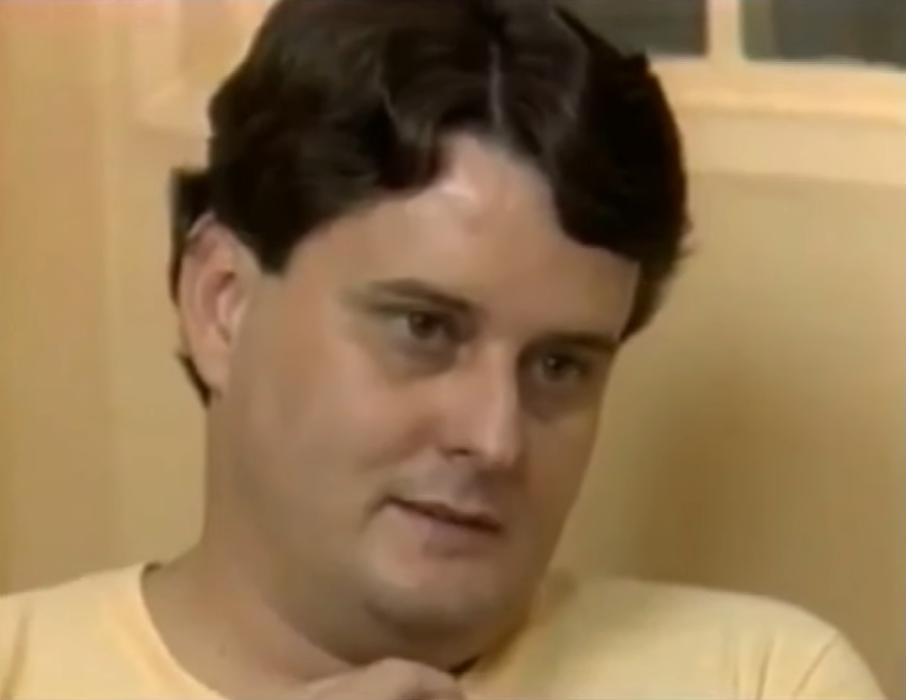 A person with short dark hair looking slightly down, wearing a light shirt, in a candid setting