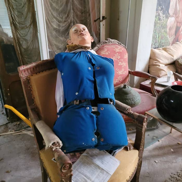 Half a mannequin sitting connected  a worn-out seat  successful  an eclectic, cluttered room, wearing a bluish  outfit with missing sleeves and buttons