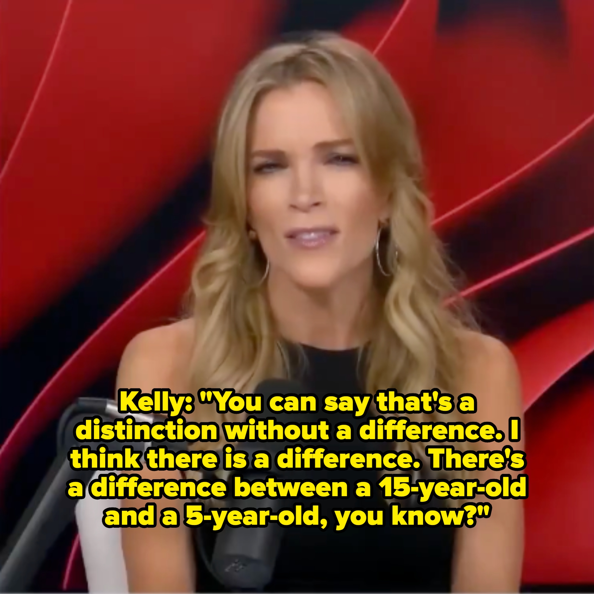 A woman with long hair speaks into a microphone, sitting in front of a red background, wearing a sleeveless top