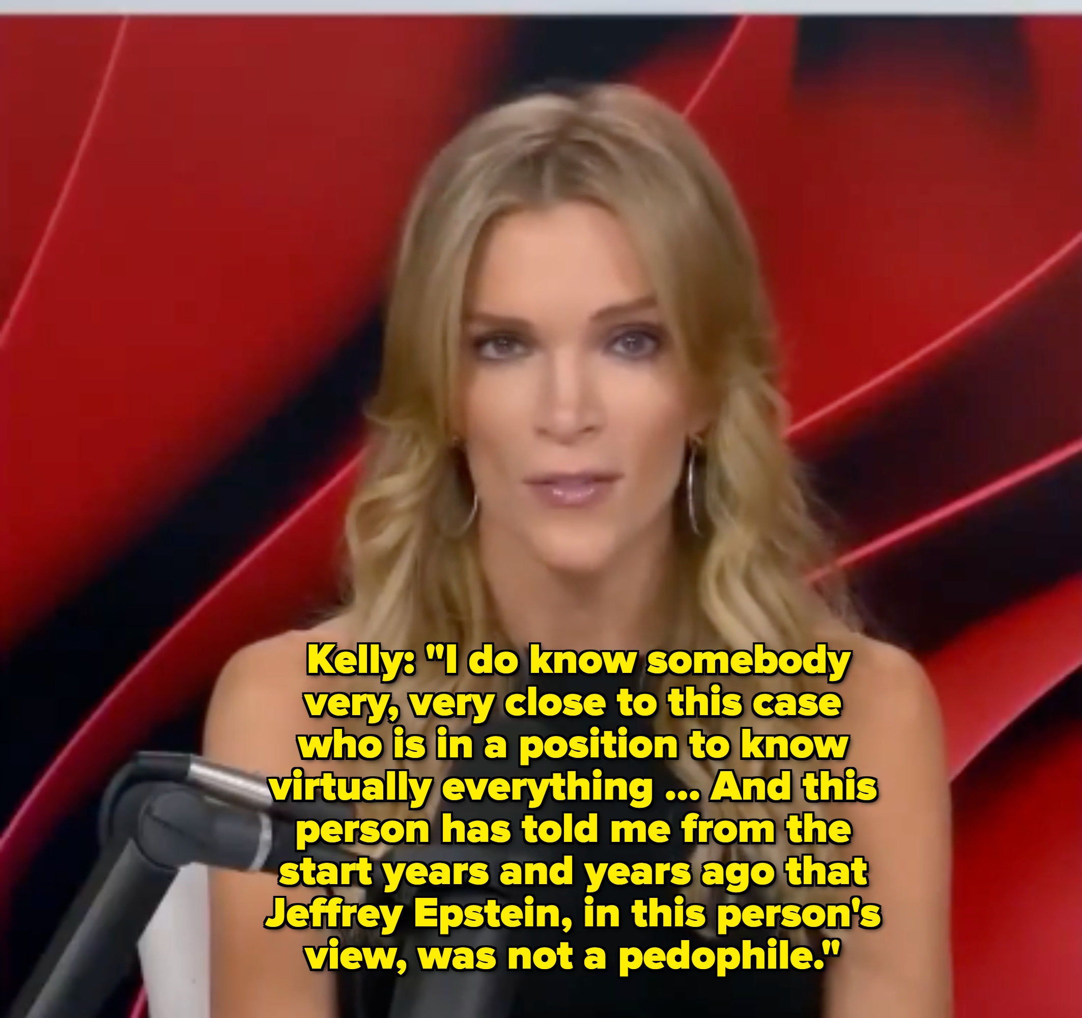 Person with long hair speaks into a microphone during a broadcast, seated against a dynamic abstract backdrop