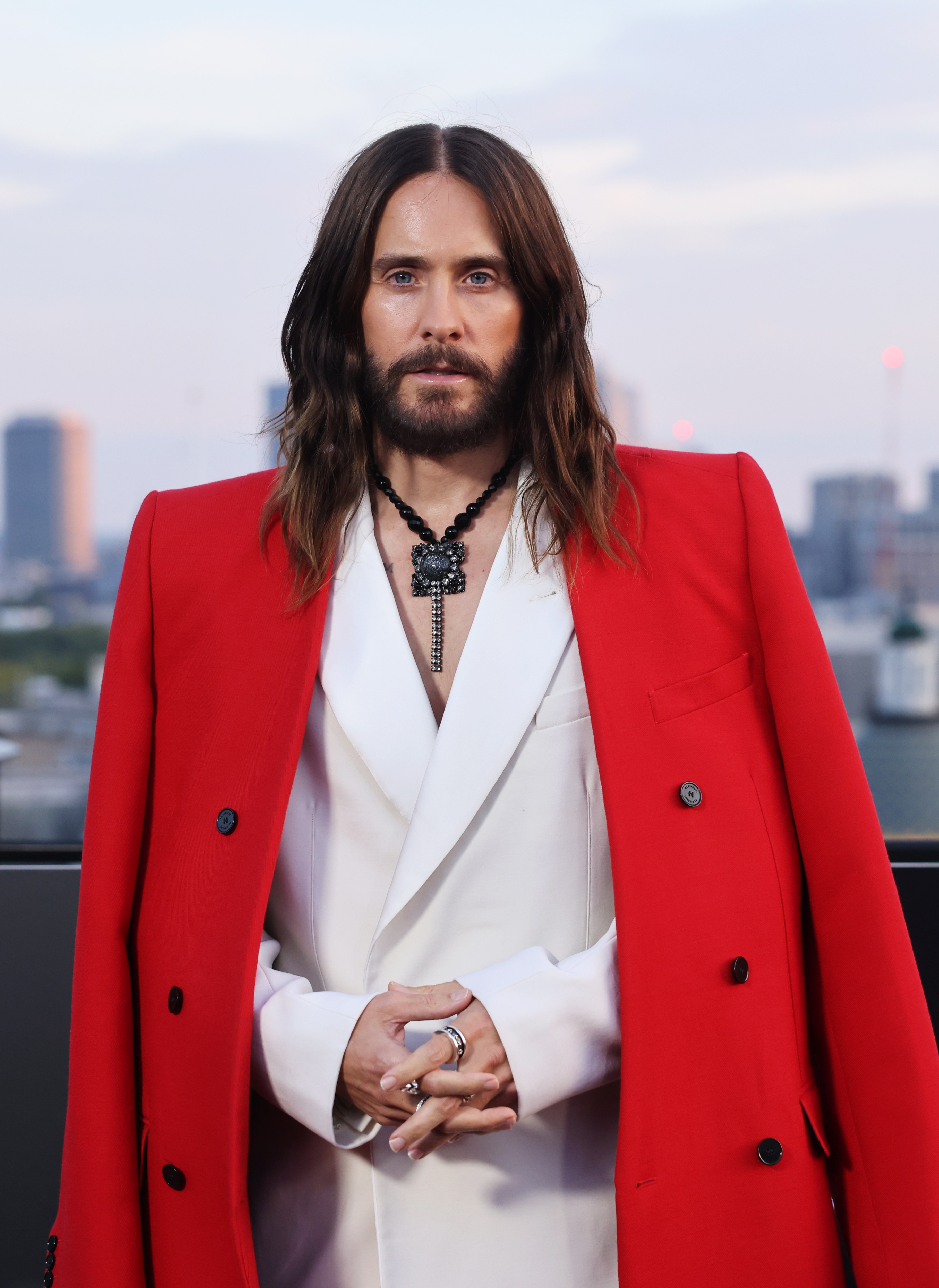 Person with agelong hairsbreadth wears a achromatic suit nether a vibrant overgarment and layered necklaces, posing connected a rooftop with a metropolis skyline successful the background