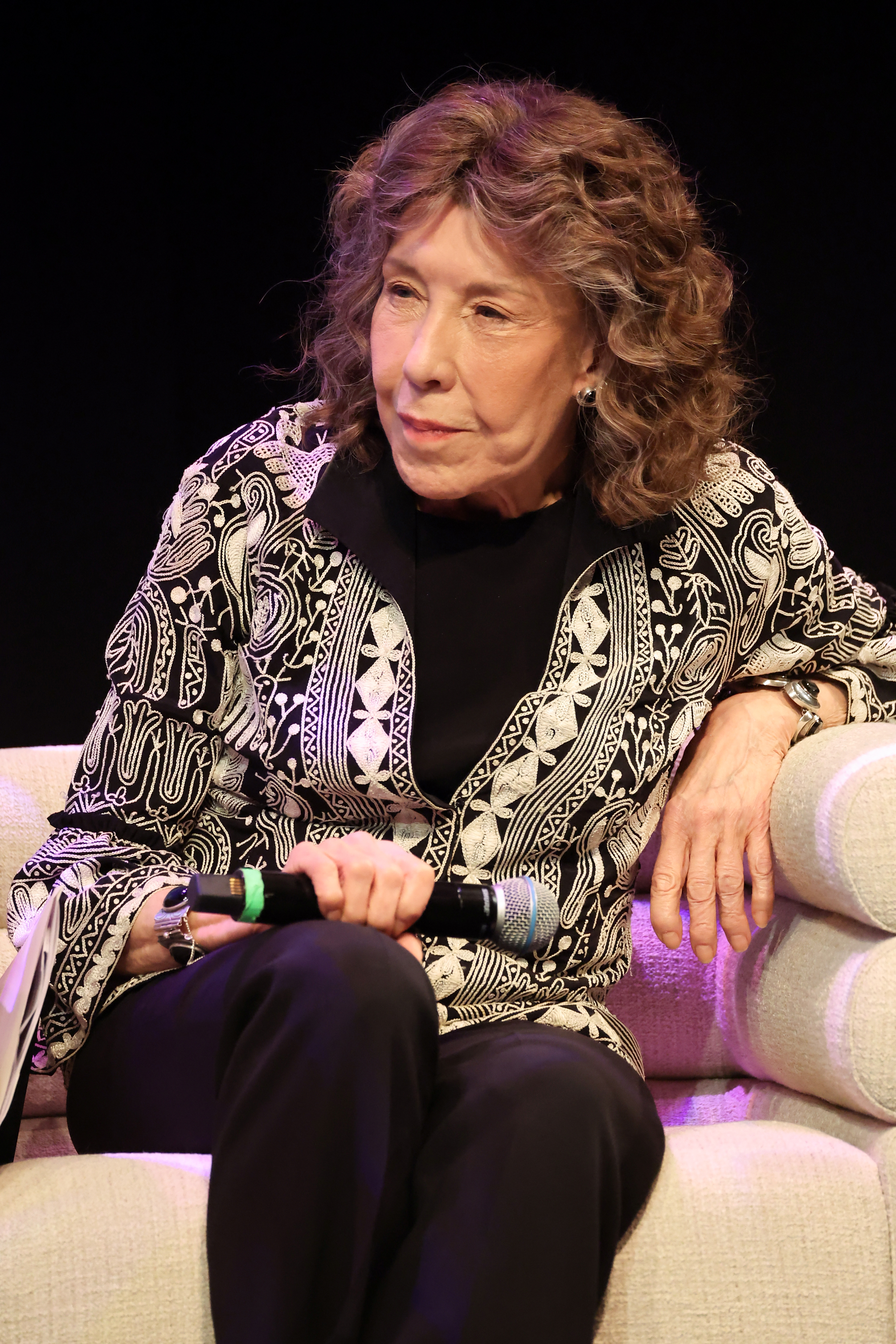 Person wearing a patterned overgarment and holding a microphone, sitting connected a pick sofa during an event