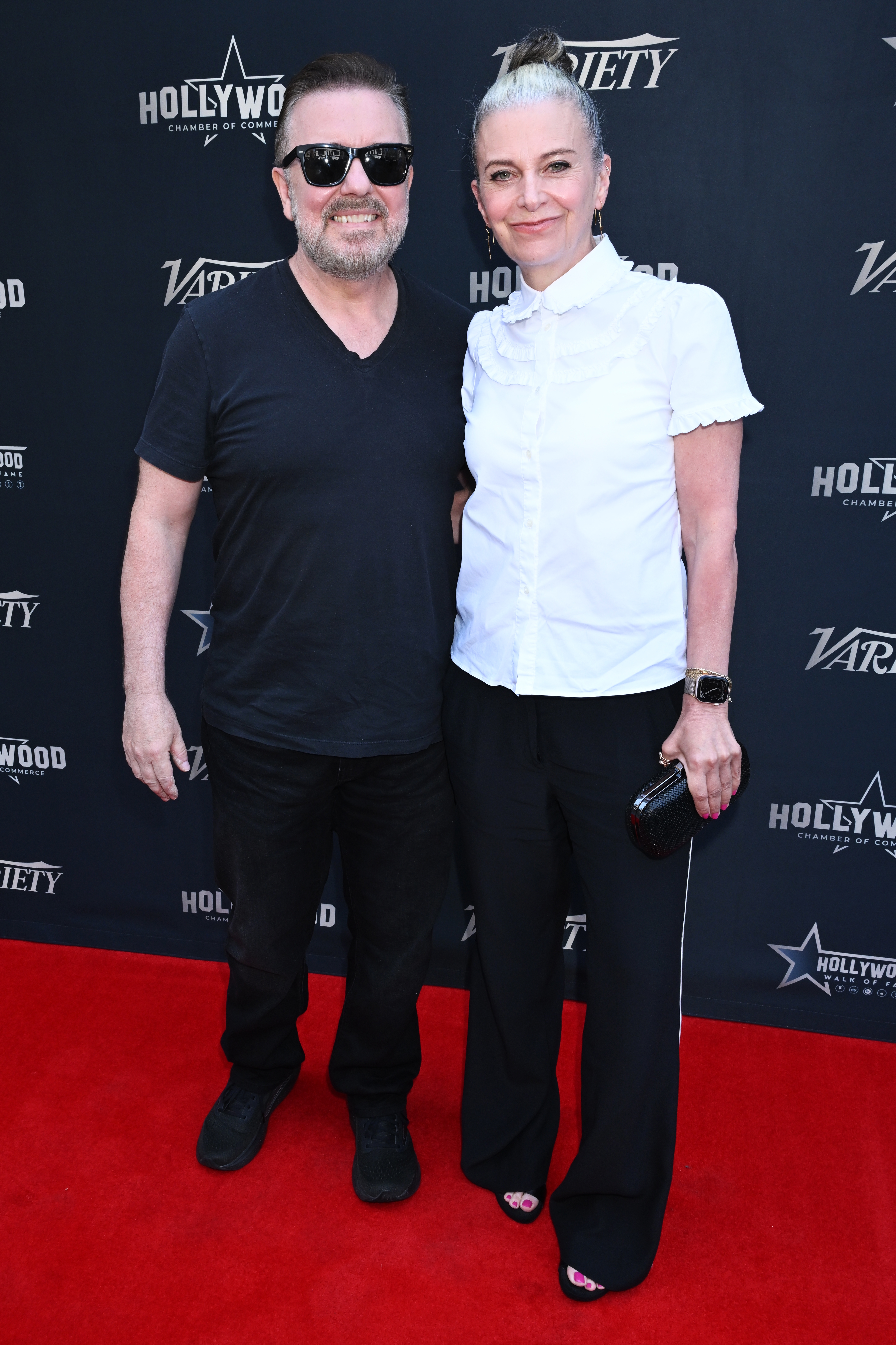Two radical posing connected a reddish carpet; 1 wears a casual achromatic outfit, the different a achromatic blouse and achromatic pants, holding a clutch