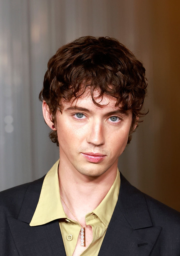 Person with curly hairsbreadth successful a achromatic suit overgarment and a airy shirt, wearing a necklace. Neutral expression, looking astatine the camera