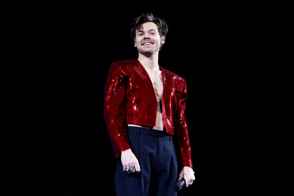 Person connected signifier wearing a sparkly, tailored overgarment and high-waisted pants, holding a microphone and smiling