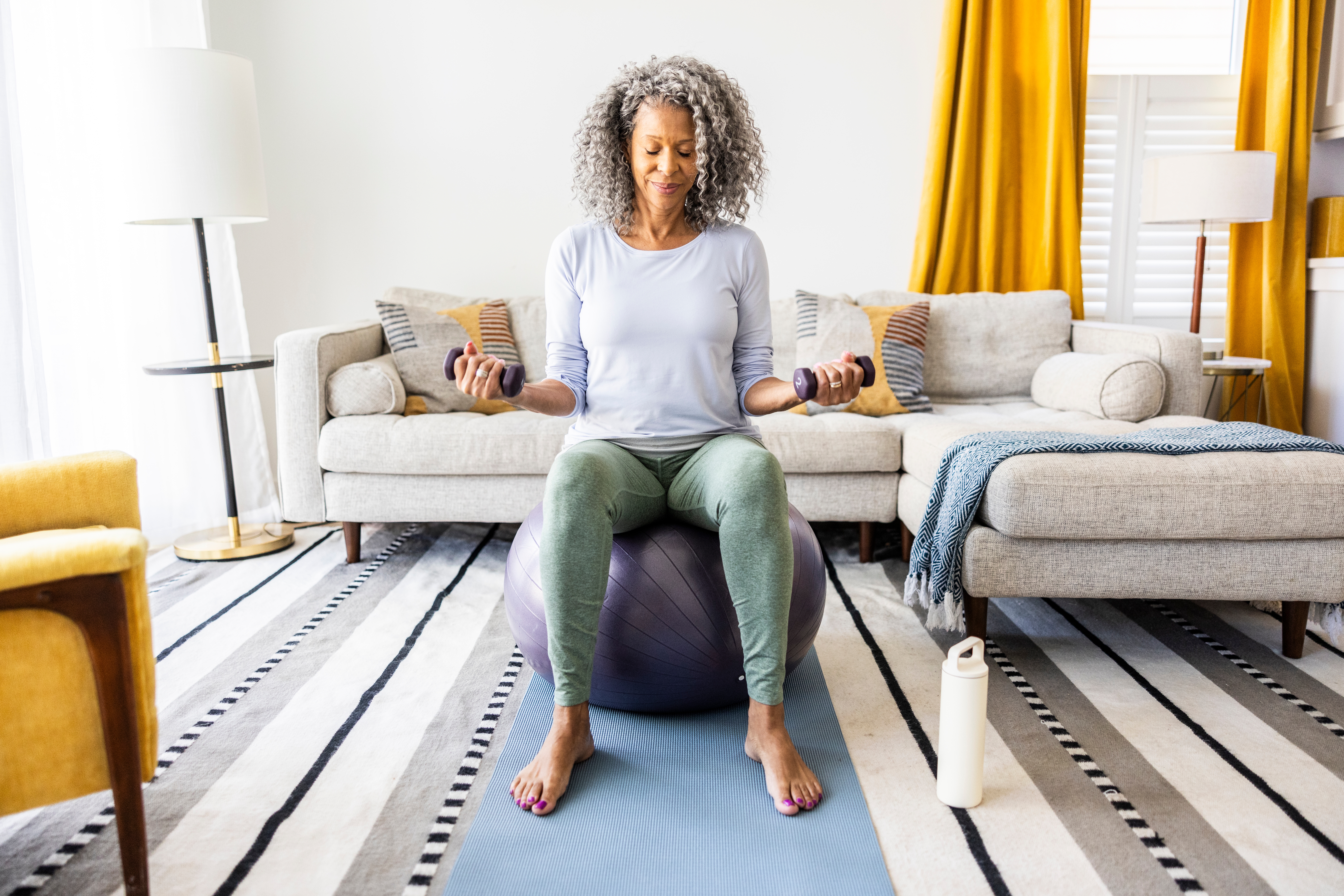 En person sitter på en treningsball og mediterer i en stue. Yogamatte og vannflaske er i nærheten