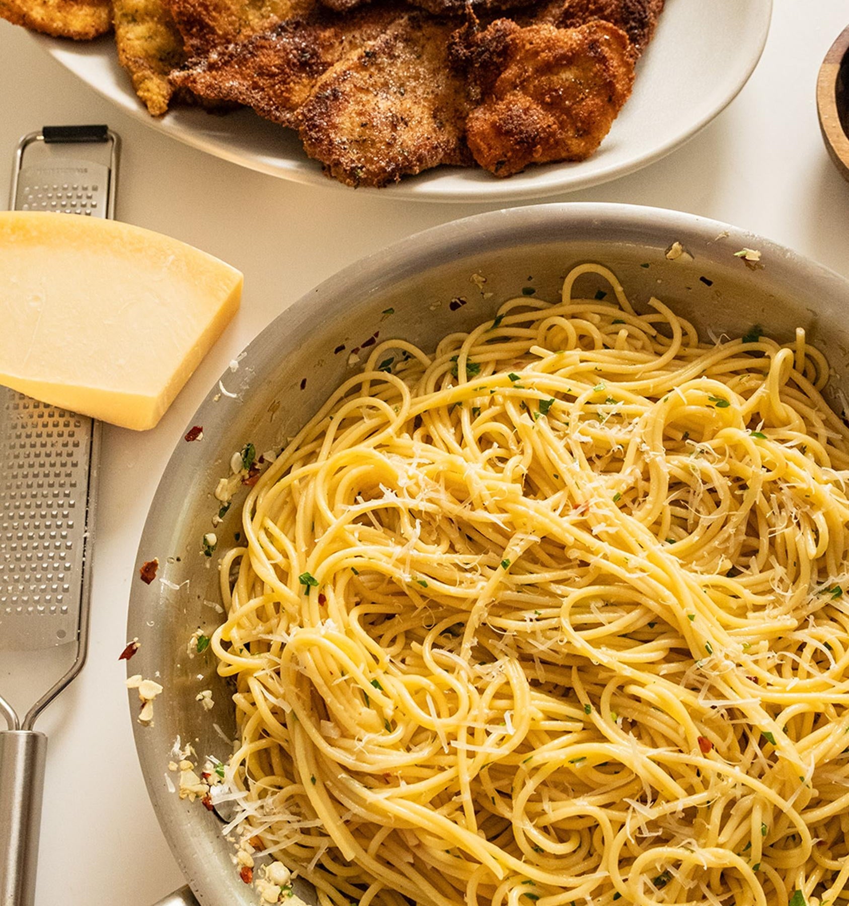 Plate of breaded, fried chicken cutlets next to a pan of seasoned spaghetti. A block of cheese and grater are to the side