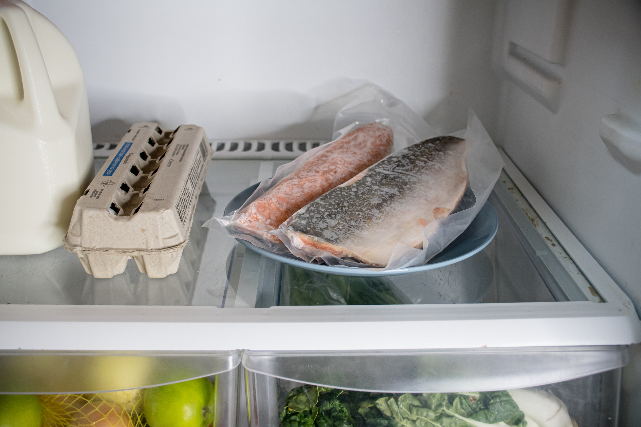 Fridge shelf with milk, eggs, and a plate containing two frozen fish fillets in a plastic bag. Below are fruits and leafy greens in a drawer
