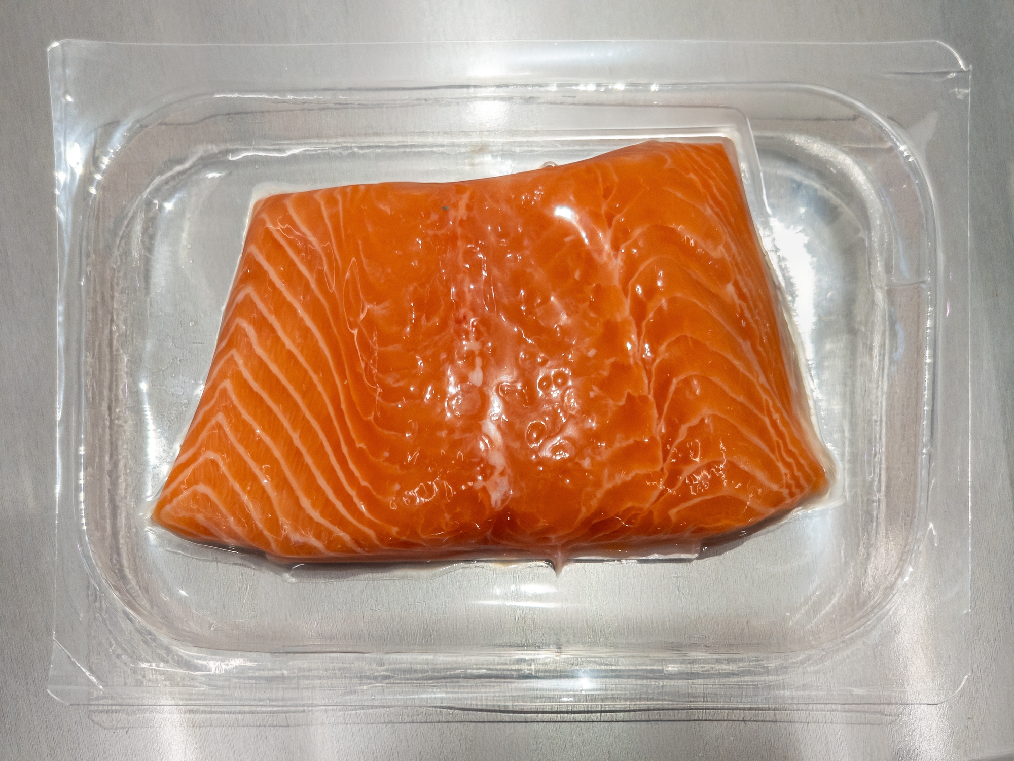 Salmon fillet in clear plastic packaging on a metal surface, showcasing rich texture and fresh appearance. Perfect for cooking or meal prep