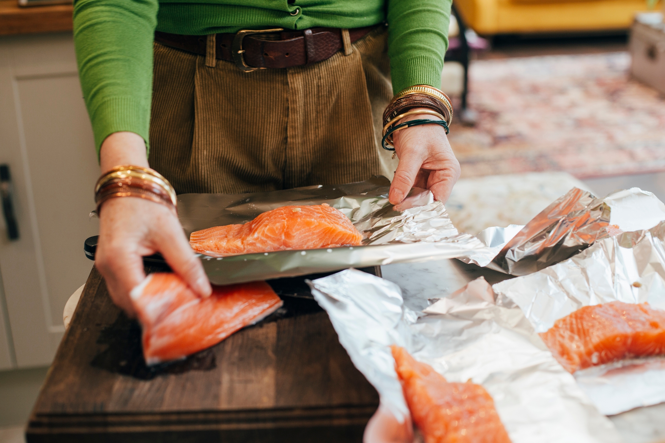 Person preparing salmon fillets on foil in a kitchen setting, wearing corduroy pants and bracelets, highlighting food preparation technique