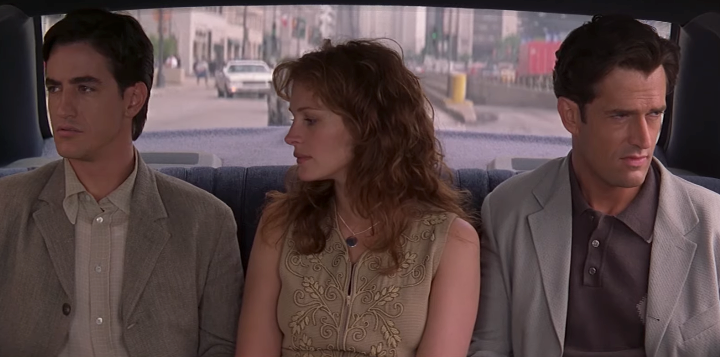 Three people sit in a car, looking in different directions with serious expressions. The woman wears an embellished top; the men wear suits