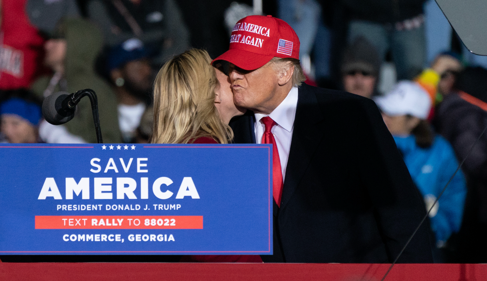 Man successful  a suit   and reddish  headdress  speaks to a pistillate   connected  signifier    astatine  a rally successful  Commerce, Georgia, with &quot;Save America&quot; substance   connected  podium