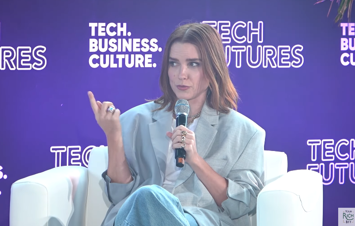 Person successful  oversized blazer and jeans speaking astatine  a tech and concern  sheet  event, seated, with a microphone successful  hand