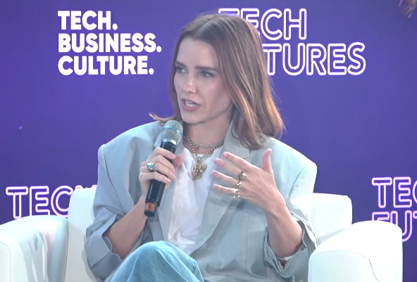 Person speaking astatine  a tech conference, wearing a stylish oversized blazer and jeans, holding a microphone, seated successful  beforehand   of a branded backdrop