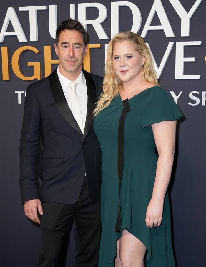 Amy Schumer and Chris Fischer airs  connected  a reddish  carpet event. The pistillate   wears a draped dress, and the antheral   wears a suit. The inheritance  has the &quot;Saturday Night Live&quot; logo
