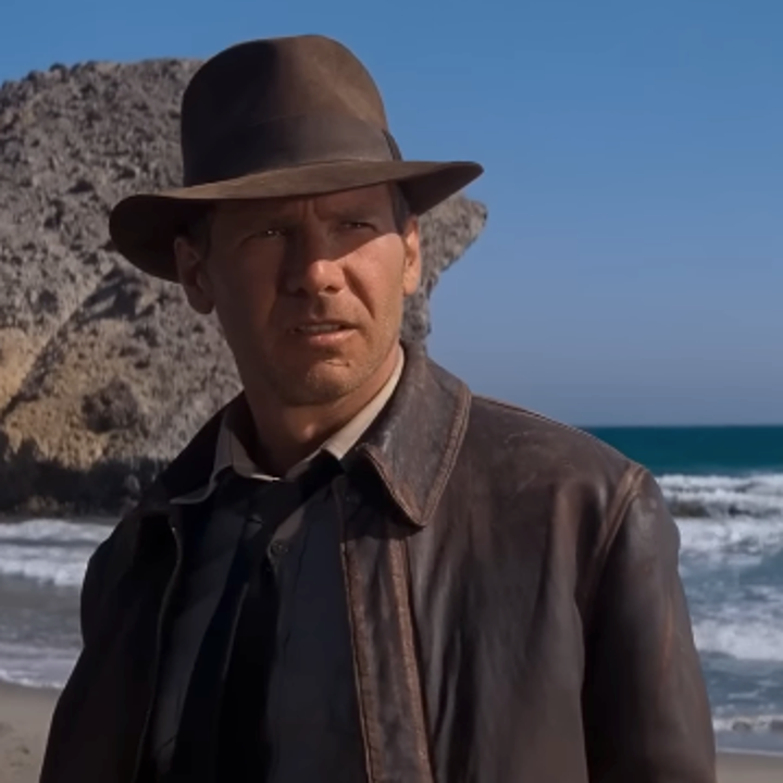 A person wearing a fedora and leather jacket stands on a beach, facing the camera with a rocky cliff in the background