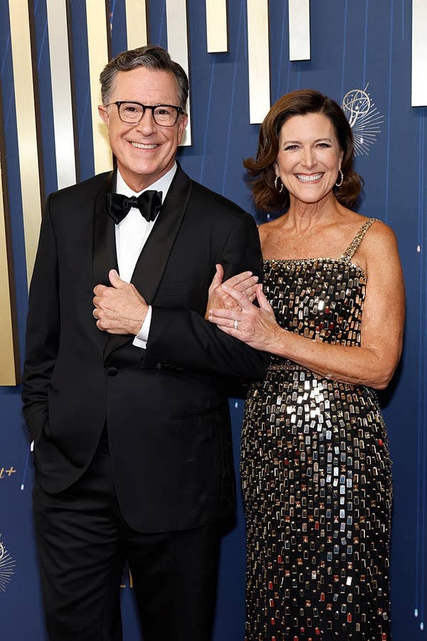 Two people on a red carpet. The person on the left wears a tuxedo; the person on the right wears a sequined, patterned gown. They’re smiling