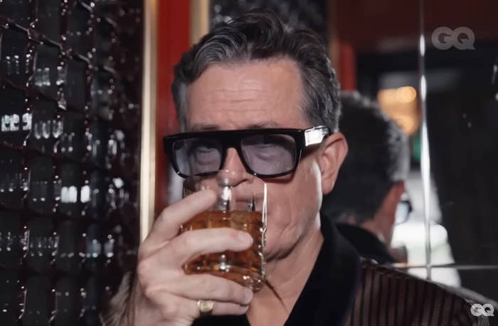 Person in sunglasses holding a glass of drink, wearing a striped robe-style outfit, standing near a reflective surface