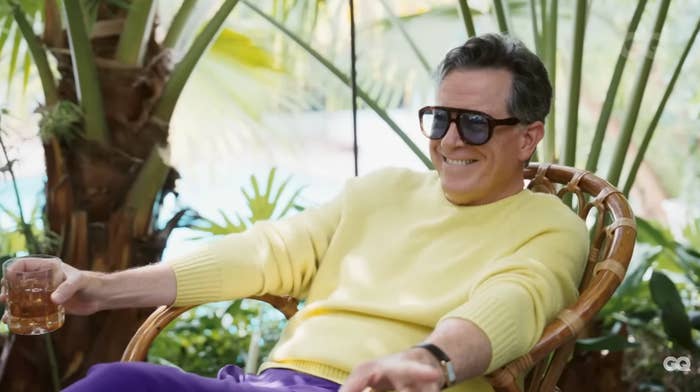A person wearing sunglasses and a casual, light sweater relaxes in a wicker chair, holding a drink, surrounded by tropical plants