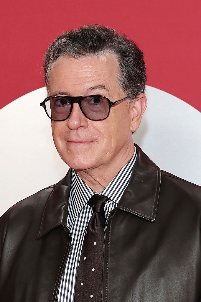 Person with sunglasses and a striped shirt under a brown leather jacket on a red carpet