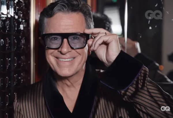 Man in a striped robe and oversized glasses smiles, stands indoors against a mirrored wall