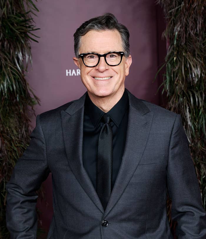 Person in a formal suit with glasses, smiling for the camera at a celebrity event. Background features greenery and a red carpet backdrop