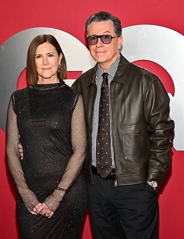 Two people pose on a red carpet. One wears a fitted dress with mesh sleeves, the other a leather jacket over a shirt and tie with polka dots