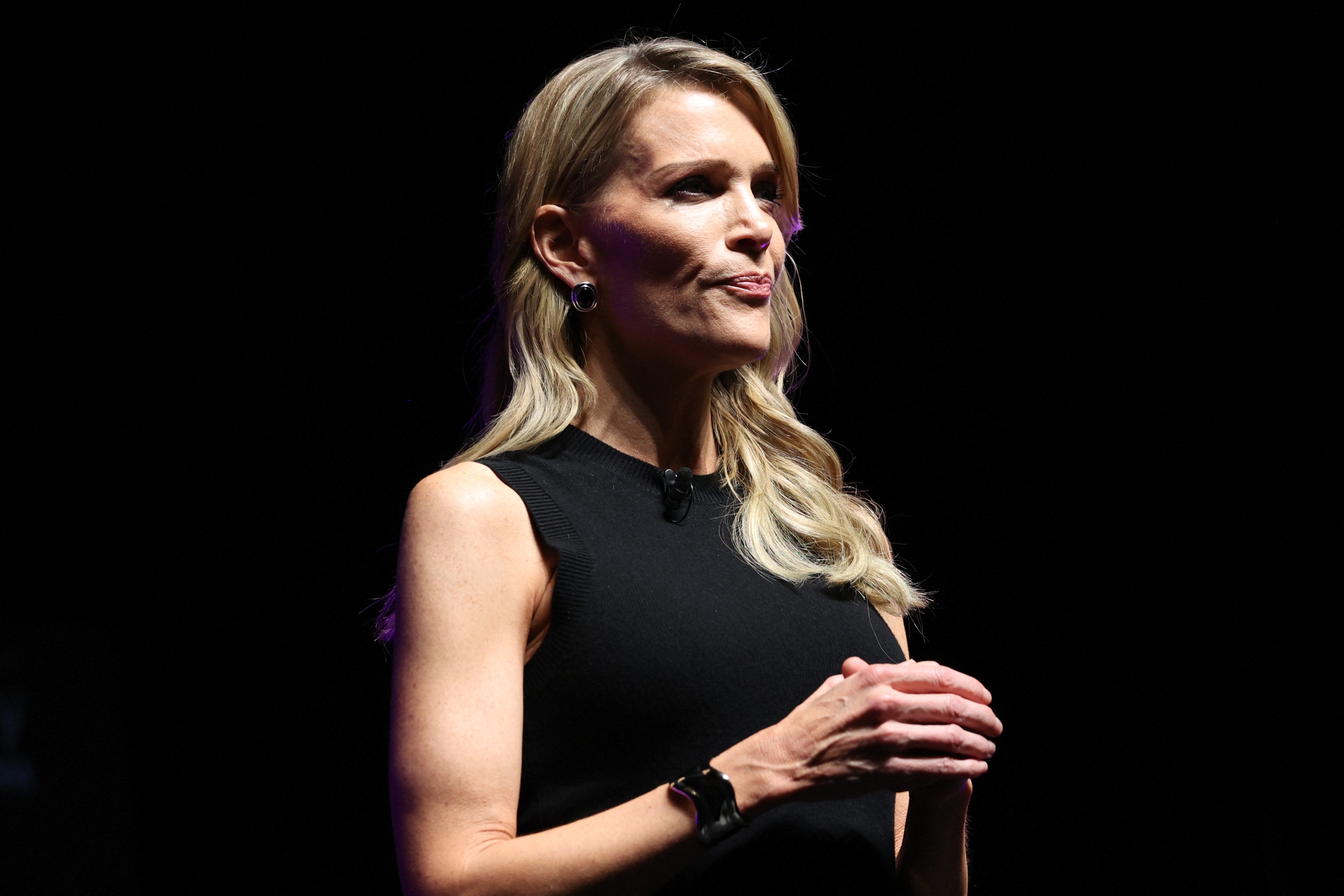 A idiosyncratic   stands connected  stage, wearing a sleeveless achromatic  dress, speaking into a microphone. Their look  is focused and attentive