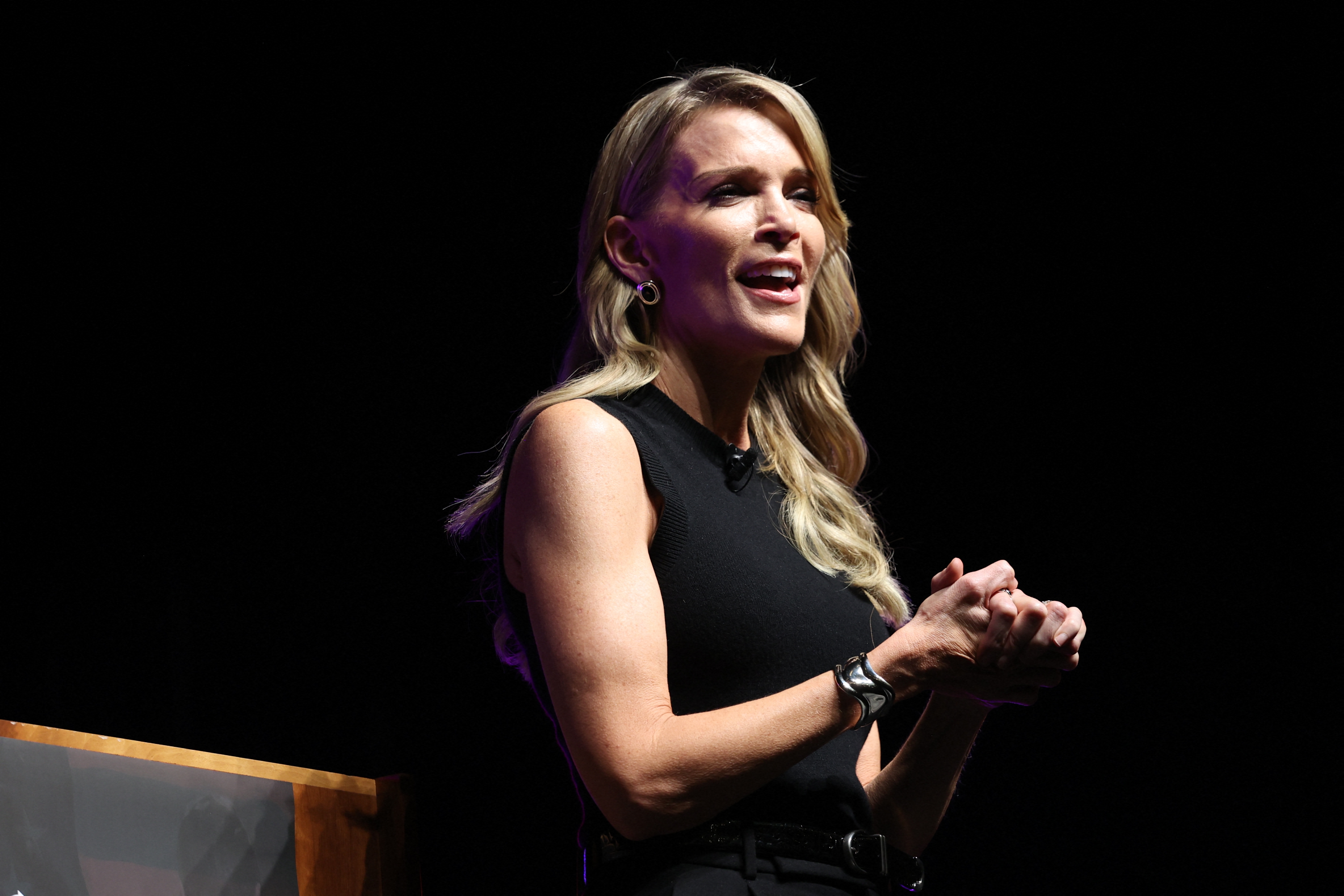 A speaker with long, wavy hair and hoop earrings addresses an audience, wearing a sleeveless top and watch, on a stage