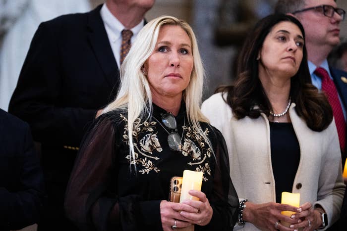 Marjorie Taylor Greene holding a candle wears a achromatic  embroidered blouse astatine  an indoor event. She stands among others, attentively watching something