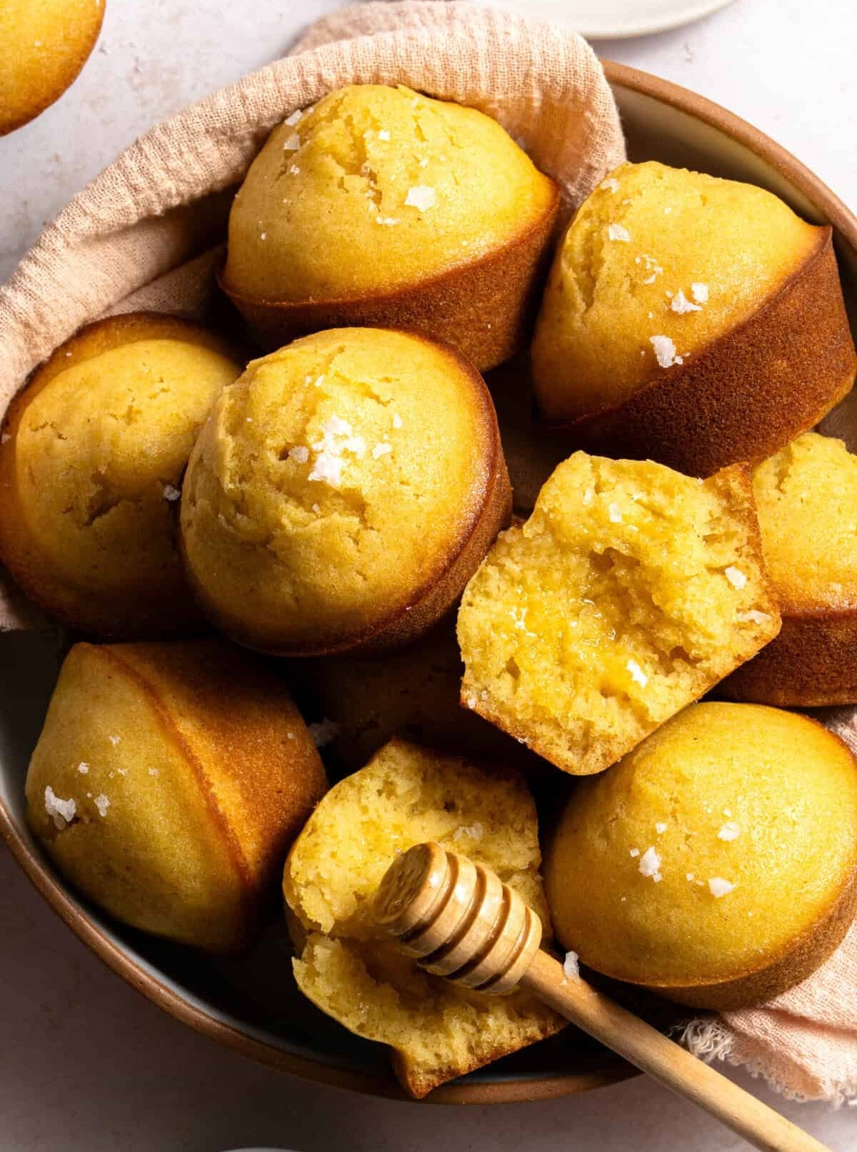 A bowl of freshly baked cornbread muffins, with one muffin split open showing its texture, accompanied by a honey dipper