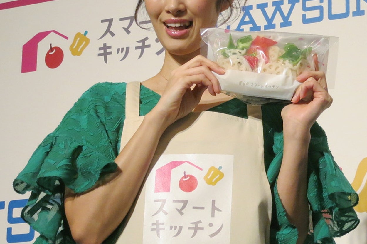 女性がグリーンのフリル袖の服とエプロンを着用し、パック入りの食品を持っていて、イベントでプロモーション活動をしています。