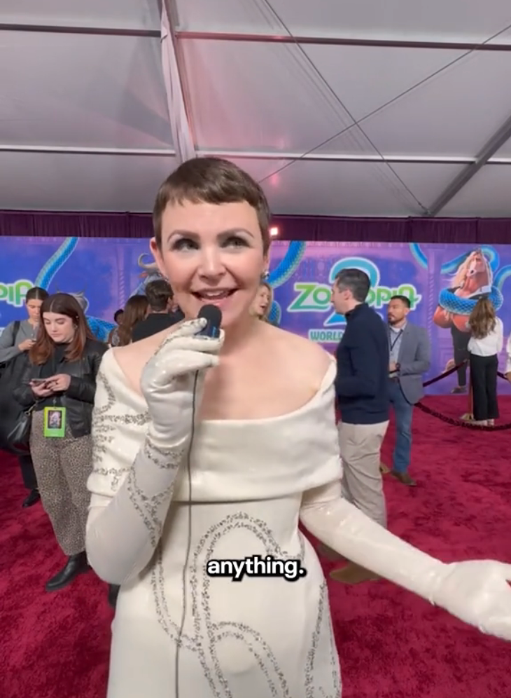 Person successful off-shoulder achromatic formal with intricate plan and gloves, speaking into a microphone connected a reddish carpet astatine a premiere event