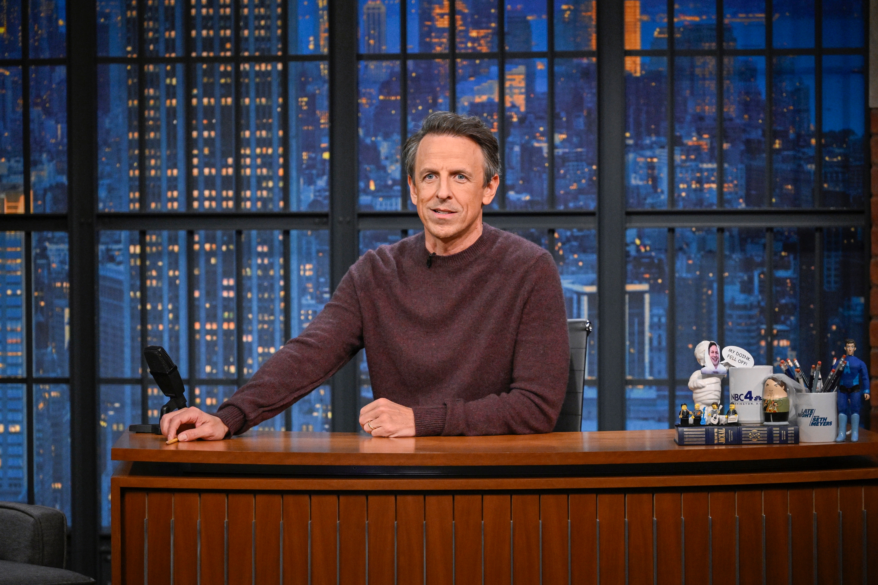 Talk amusement big seated astatine table successful beforehand of ample model with cityscape backdrop, smiling, wearing casual sweater, assorted table items visible