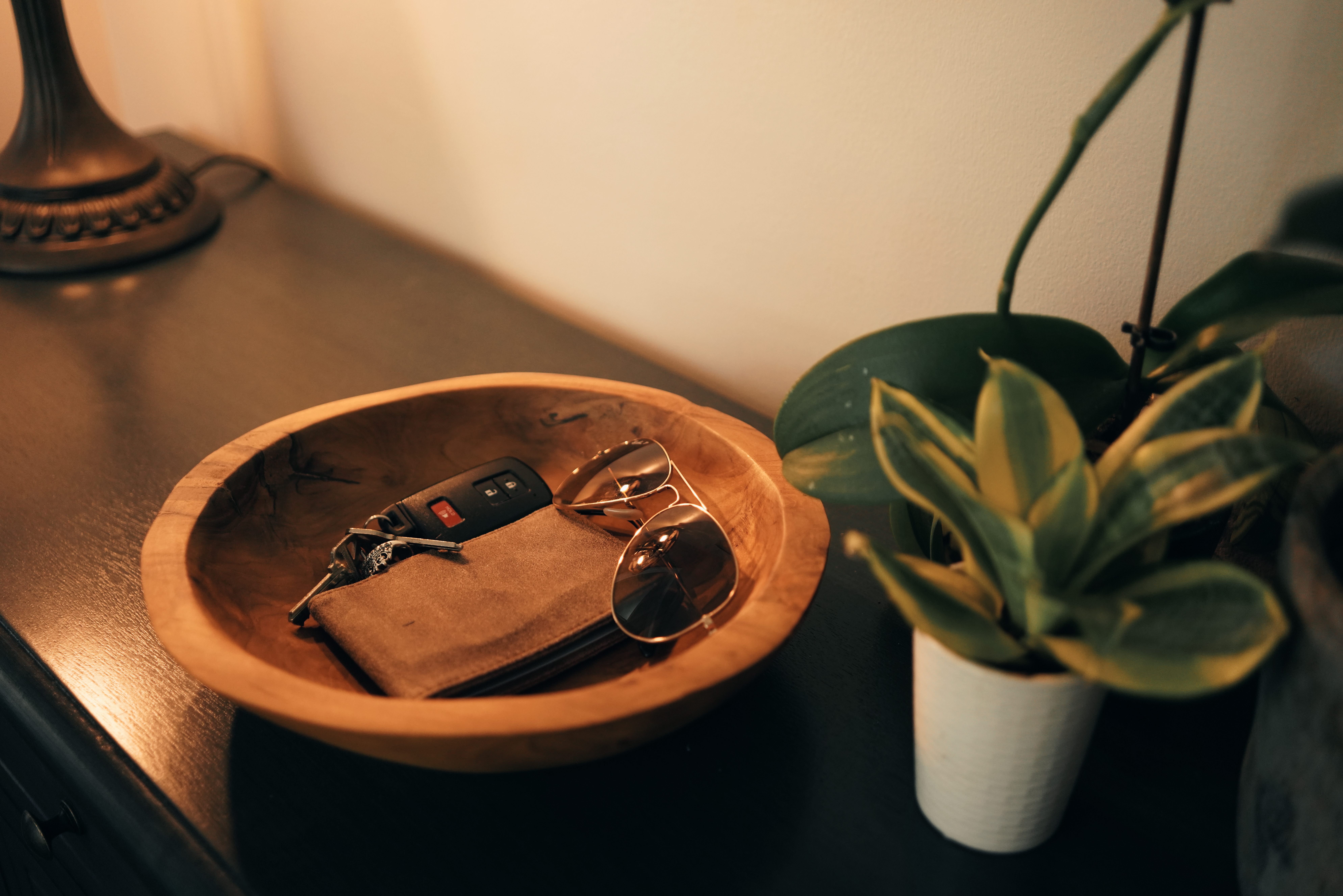 En treskål inneholder solbriller, en lommebok og bilnøkler. En liten potteplante står ved siden av på et mørkt bord