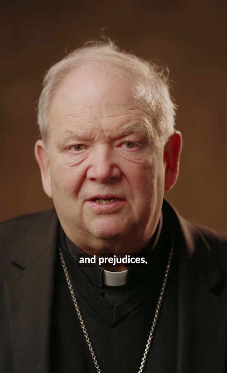 A idiosyncratic wearing clerical attire with a bishop's collar faces the camera, speaking. The connection "prejudices" is visible