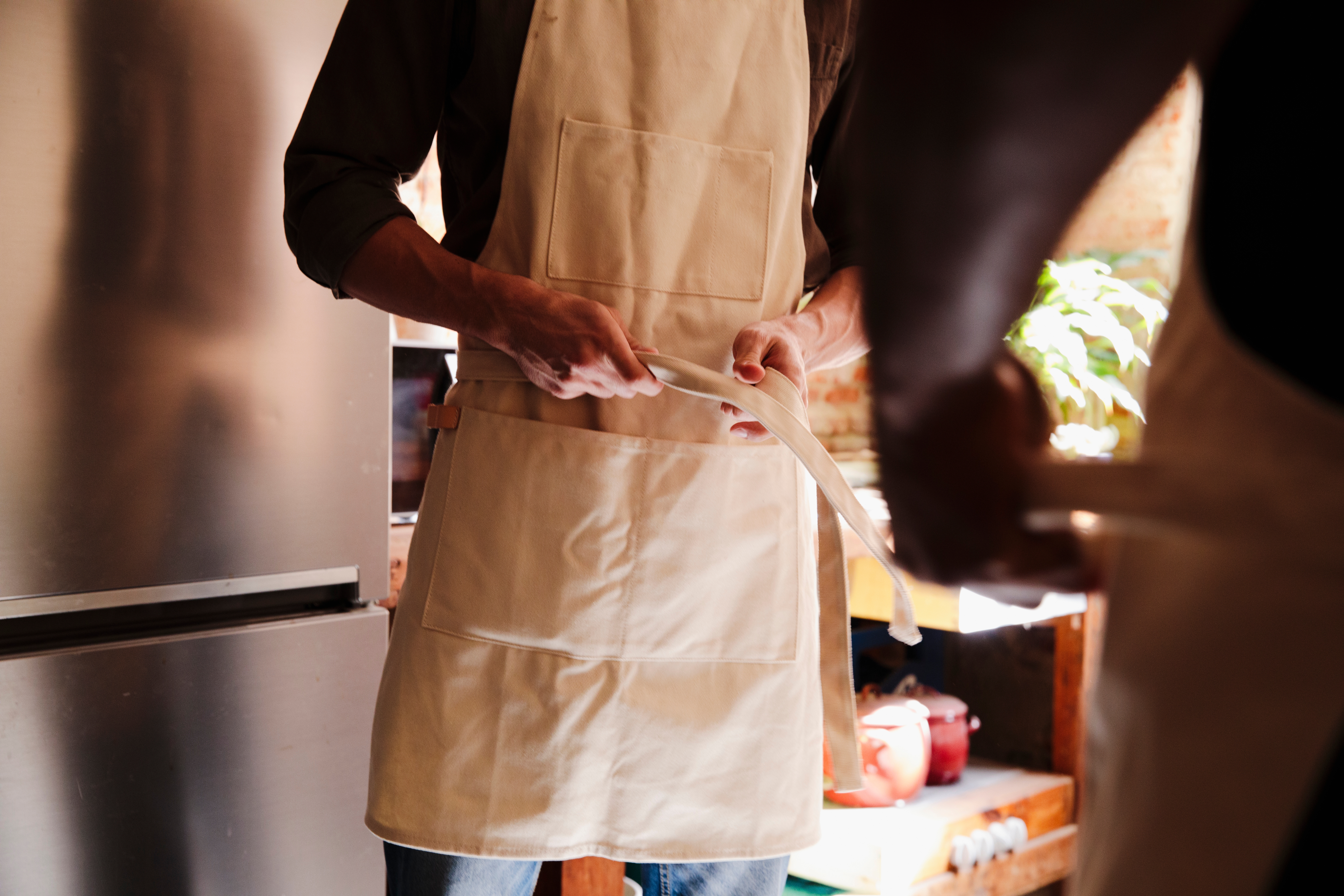 To personer på et kjøkken, iført forkle, tilbereder mat sammen. Den ene holder et redskap