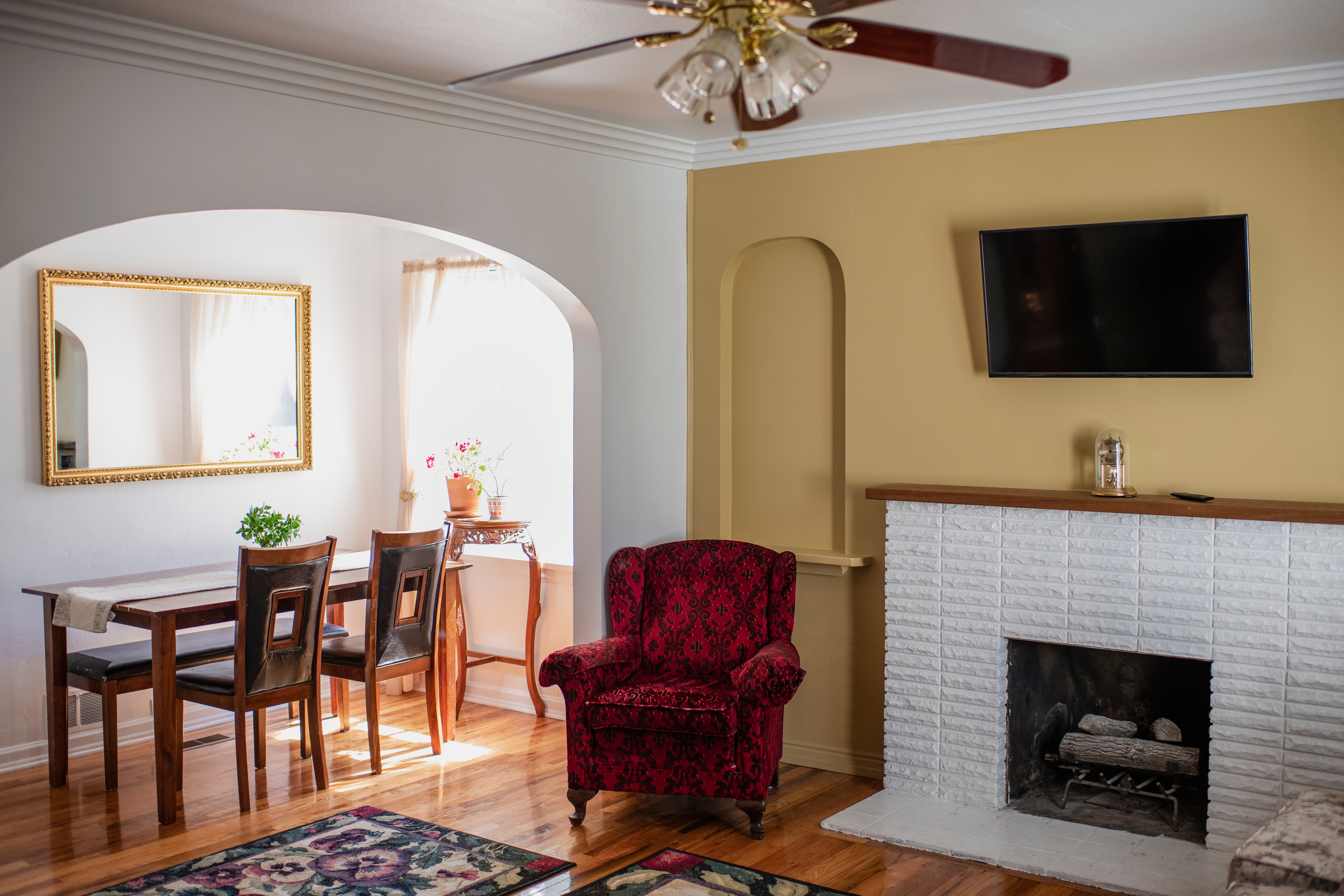 Stue med rødmønstret lenestol, spisebord, vegghengt TV og et dekorativt speil over et trebord med planter