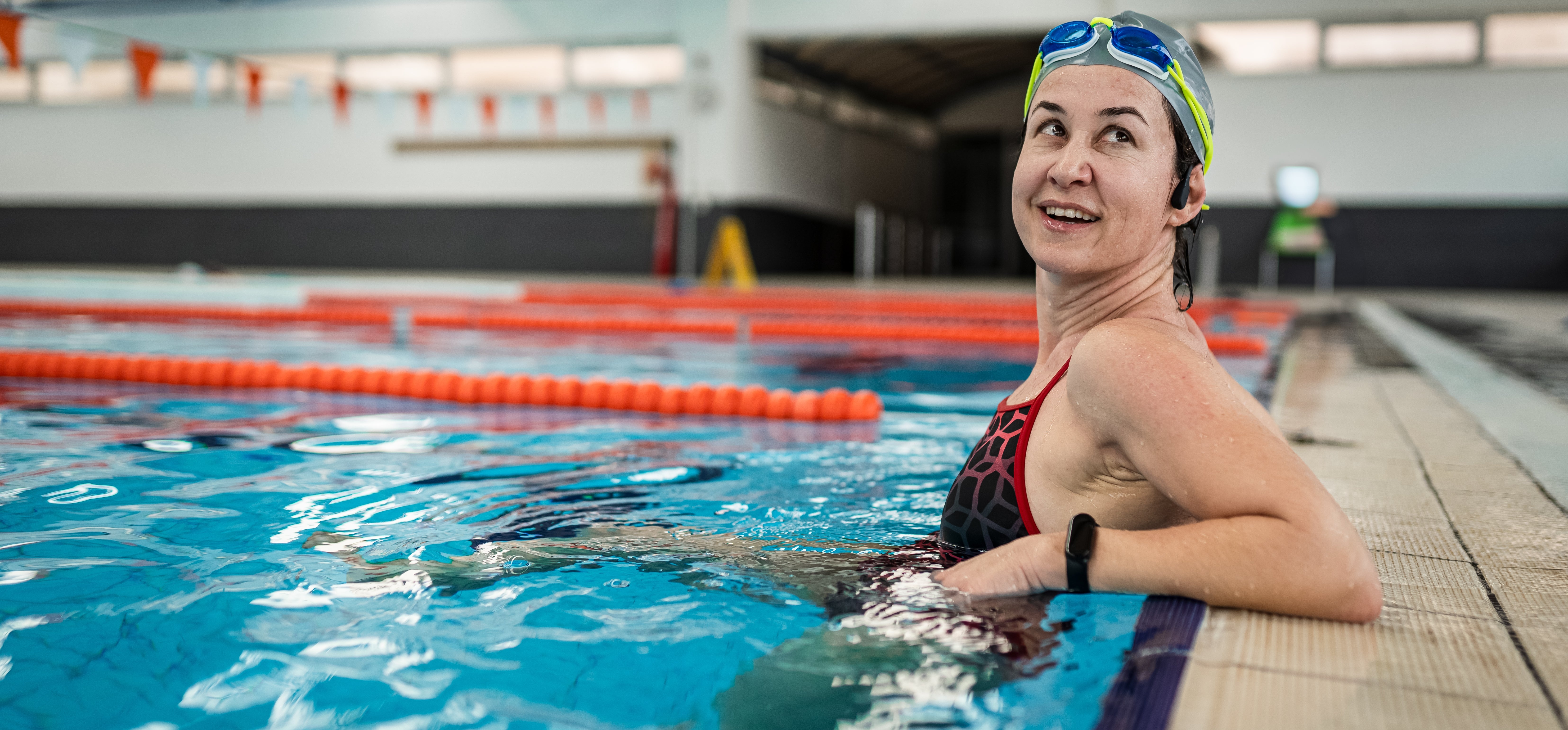Person med badehette og briller slapper av ved kanten av et innendørsbasseng, ser tilbake og smiler