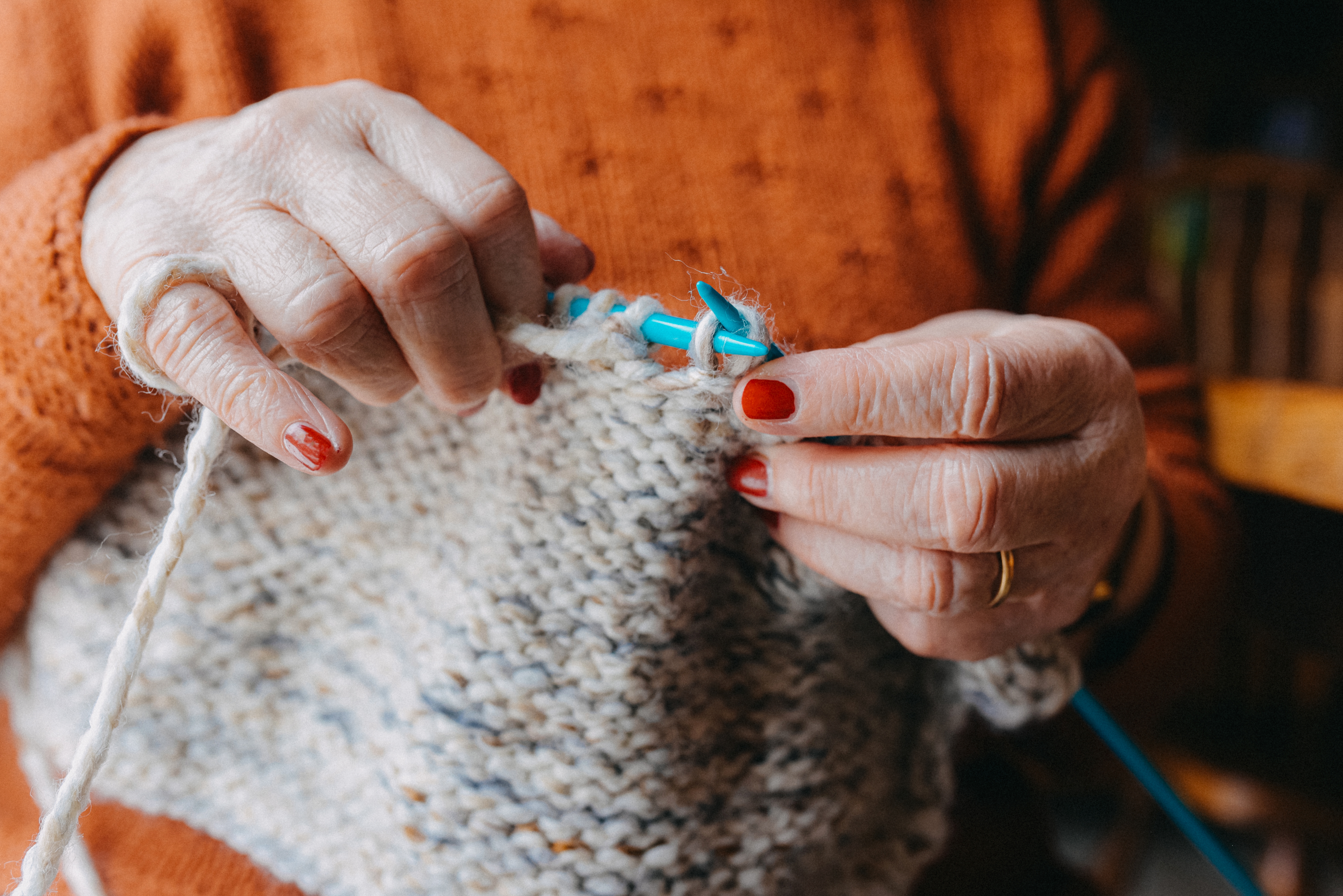 Eldre persons hender som strikker med beige garn, iført en oransje strukturgenser og en ring på en finger