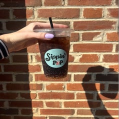 Someone holding an iced drink in a plastic cup with a branded sticker against a brick wall
