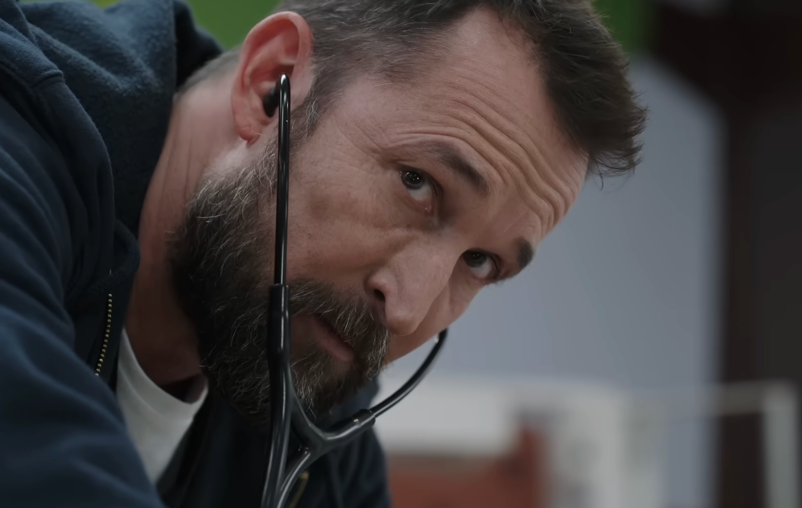 A antheral   with a beard wearing casual covering  looks intently, holding a stethoscope