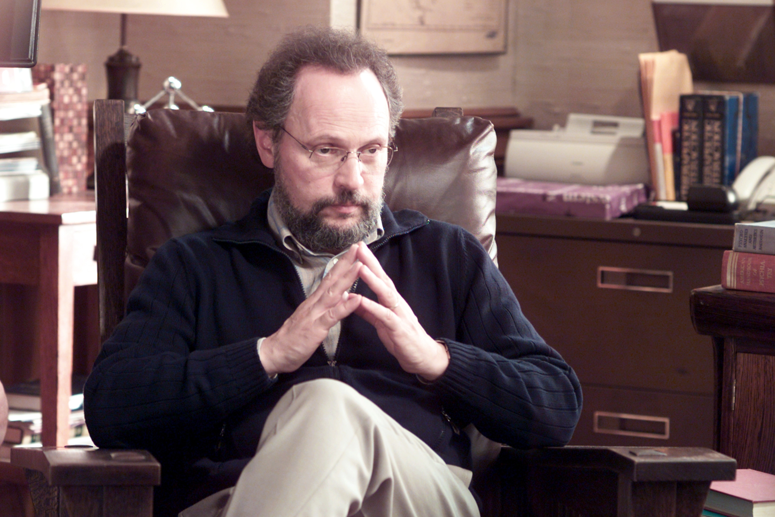 Person som sitter i en lenestol, hender som danner en trekant, virker tankefull i en studiesetting med bøker og en skriver i bakgrunnen