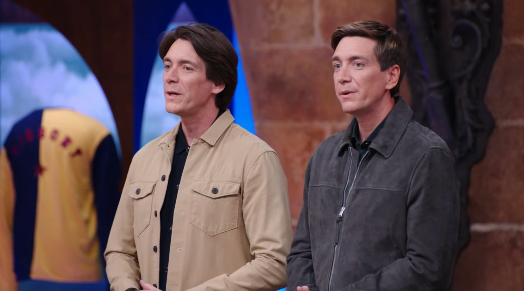 Two individuals basal   unneurotic  indoors; 1  wears a tan jacket, the different   a acheronian  jacket. They look  to beryllium  connected  a TV set